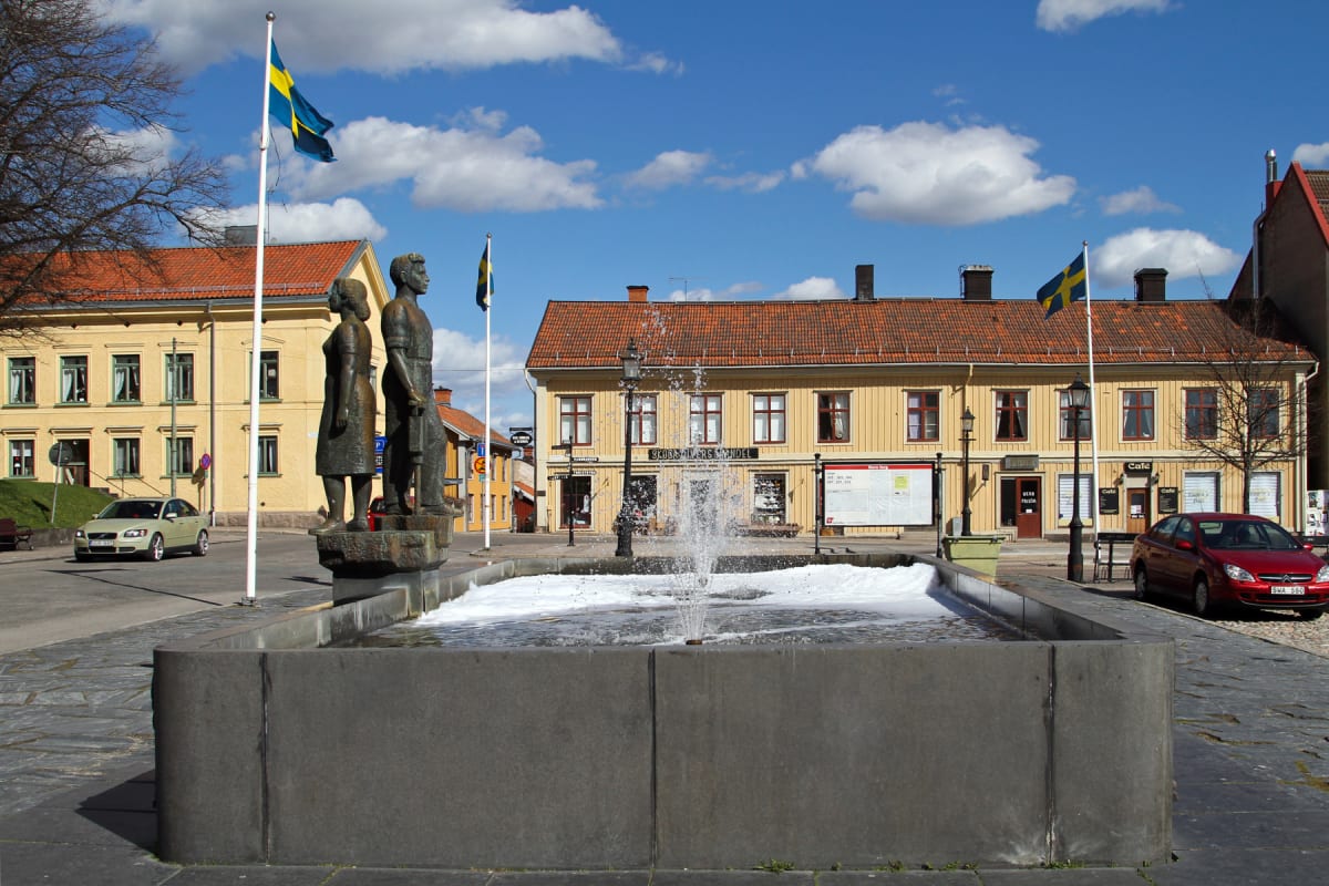 En bild av ett torg med en fontän och en skulptur föreställande två personer. I bakgrunden syns flera gula byggnader med röda tak, en flaggstång med en svensk flagga och vita moln på en blå himmel. Två bilar är parkerade eller kör på torget.