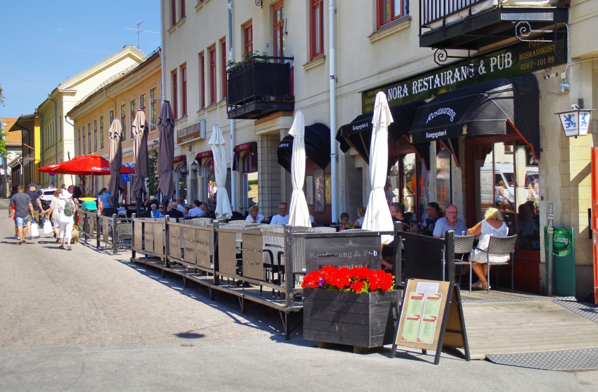 En gata med uteserveringar från restauranger och pubar. Några personer sitter vid borden med parasoller utslagna. På gatan går människor och några barn är synliga. Mellan uteserveringarna och gatan står blomlådor med röda blommor. Restaurangerna är i