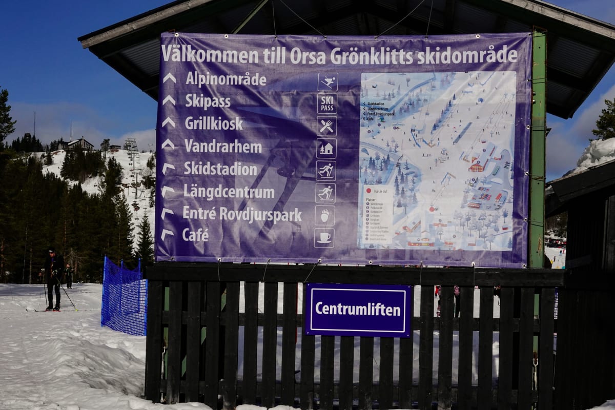 En stor lila skylt med texten ”Välkommen till Orsa Grönklitts skidområde” och en karta över skidområdet står monterad på en trävägg. Till vänster syns snö, skidspår och en person på skidor. I bakgrunden syns skog och en backe med en skidlift.