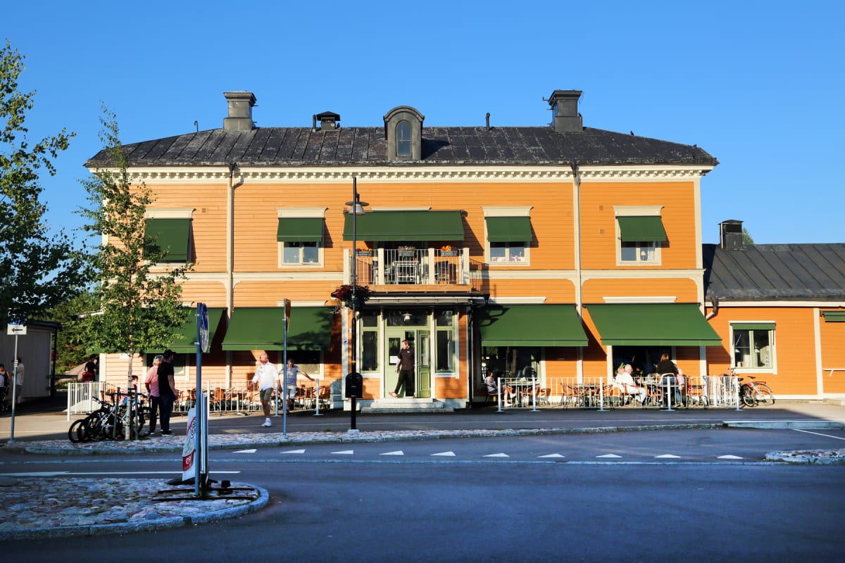 En gulorange byggnad med svart tak och gröna markiser, framför vilken en gata med en trädplanterad gatstenbeläggning löper. Utanför byggnaden sitter och står flera personer, och ett par cyklar är parkerade vid en stolpe. Himmelen är klarblå.