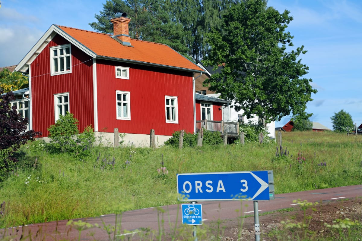 En röd stuga med tegeltak och vita fönsterkarmar står på en grönskande sluttning. En vägskyllt med texten "ORSA 3" och en cykelsymbol syns i förgrunden, delvis skymd av vegetation. Himlen är blå med några vita moln.
