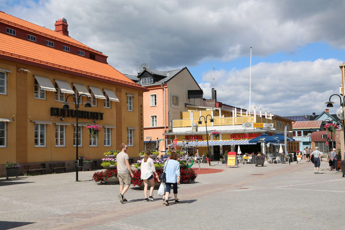 En gata eller ett torg i en stad med byggnader på båda sidor. En gul byggnad med texten "Kulturhuset" och flera fönster med markiser syns till vänster. Framför byggnaderna finns blomlådor och människor går på det stenlagda området.