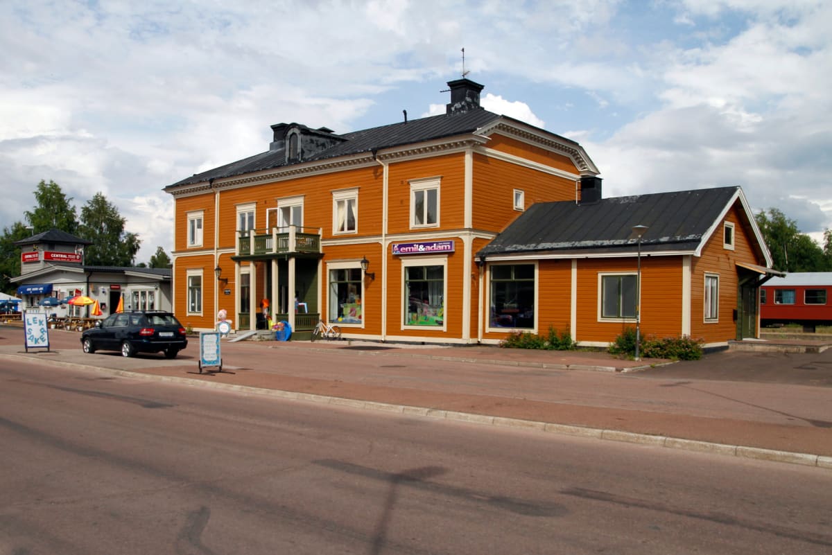 En gata framför en stor gul byggnad med vita knutar, där en svart bil står parkerad. Bakom byggnaden syns träd och en röd tågvagn. En mindre byggnad står till vänster med skyltar och parasoller. En blå cykel och reklamskyltar finns nära huvudbyggnade