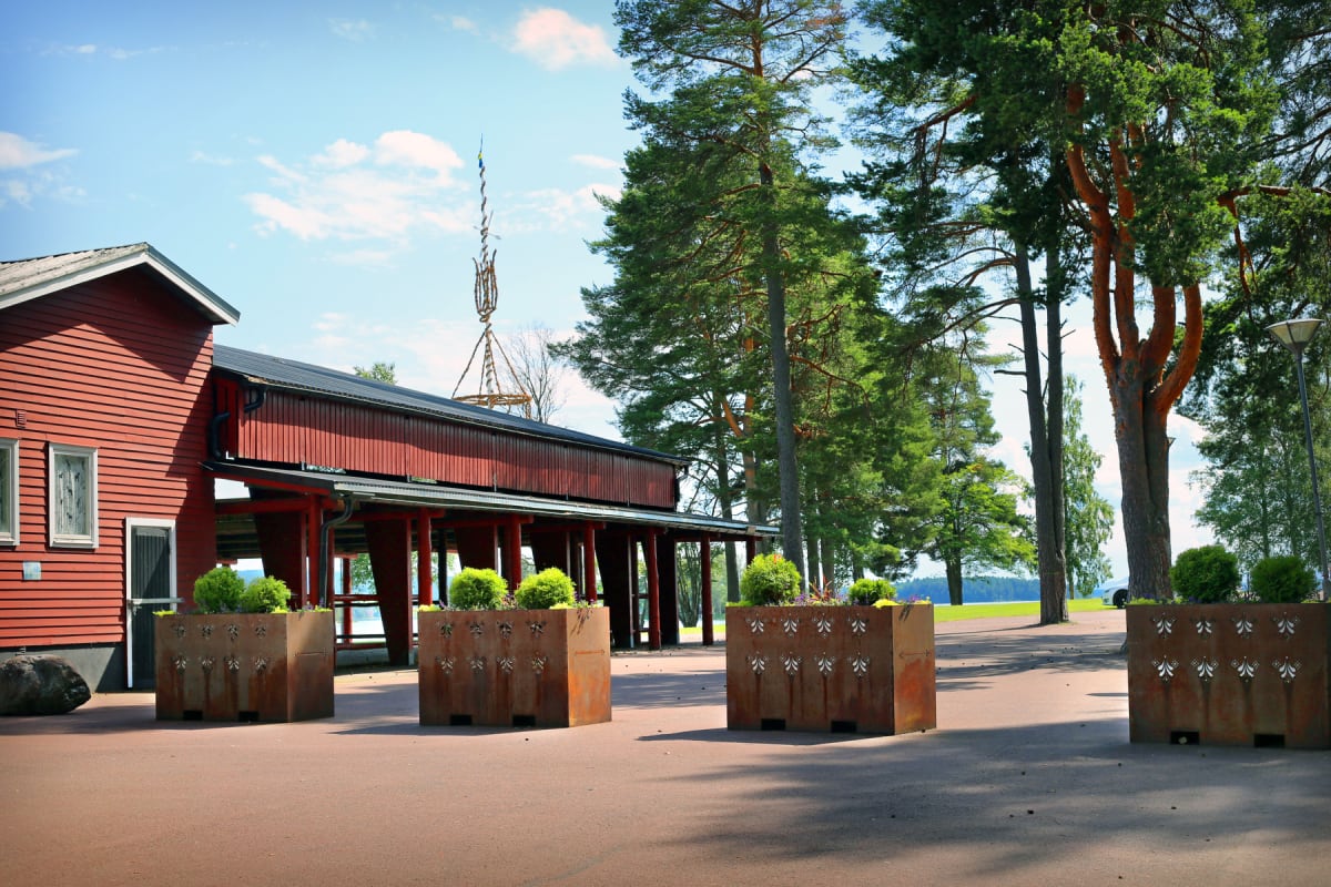 En röd byggnad med ett svart tak står till vänster, med tallar och ett vattendrag i bakgrunden. En midsommarstång syns vid hustaket. Fyra rostfärgade blomlådor med gröna växter står på det asfalterade förgrunden. Himlen är blå med sprida moln.