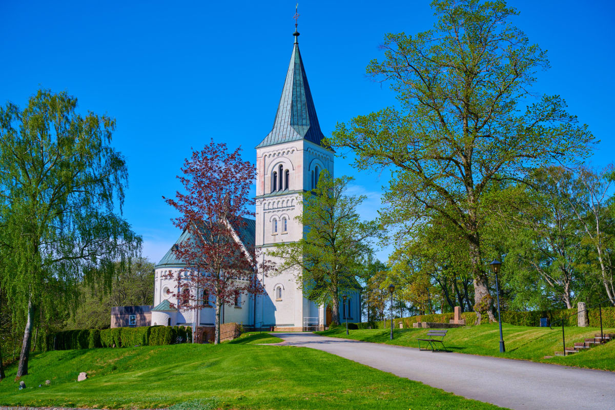 En vit kyrka med ett grått spetsigt tak som reser sig mot en klarblå himmel. Runt kyrkan finns gröna gräsmattor och lövträd med grönskande blad. En asfalterad väg leder upp mot kyrkan, och en bänk syns vid sidan av vägen. En häck löper längs gräsmatt