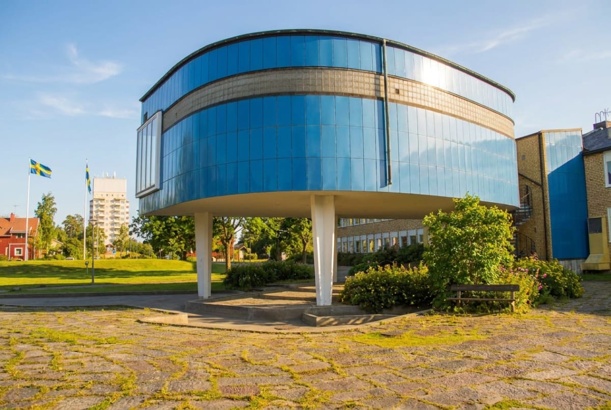 En modern byggnad med en böjd, glasfasad i blått dominerar bilden. Byggnaden vilar på pelare och under den finns buskar och marksten. I bakgrunden syns svenska flaggor på stänger, bostadshus och träd under en klarblå himmel.