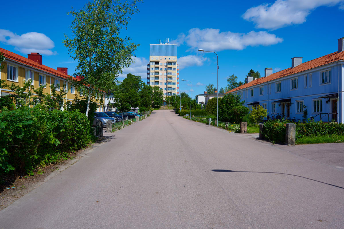 En gata med parkerade bilar och flerfamiljshus på båda sidor, under en klarblå himmel med spridda moln. Längst bort i bild syns ett högre bostadshus. Grönska som buskar och träd kantar gatan och husen.
