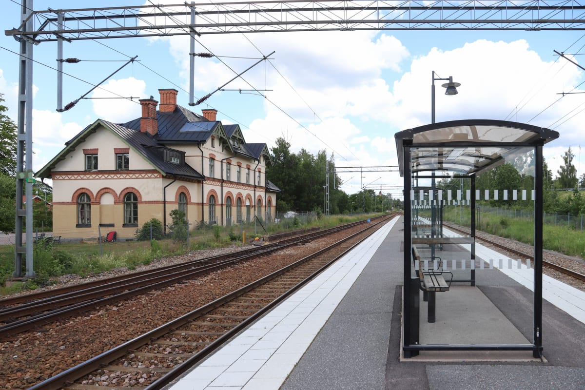 En bild som visar en tågstation med ett stationshus, spår och en perrong med väntkur. Huset är beige med bruna bågar runt fönstren och ett mörkgrönt tak. På perrongen står en modern väntkur med bänkar.