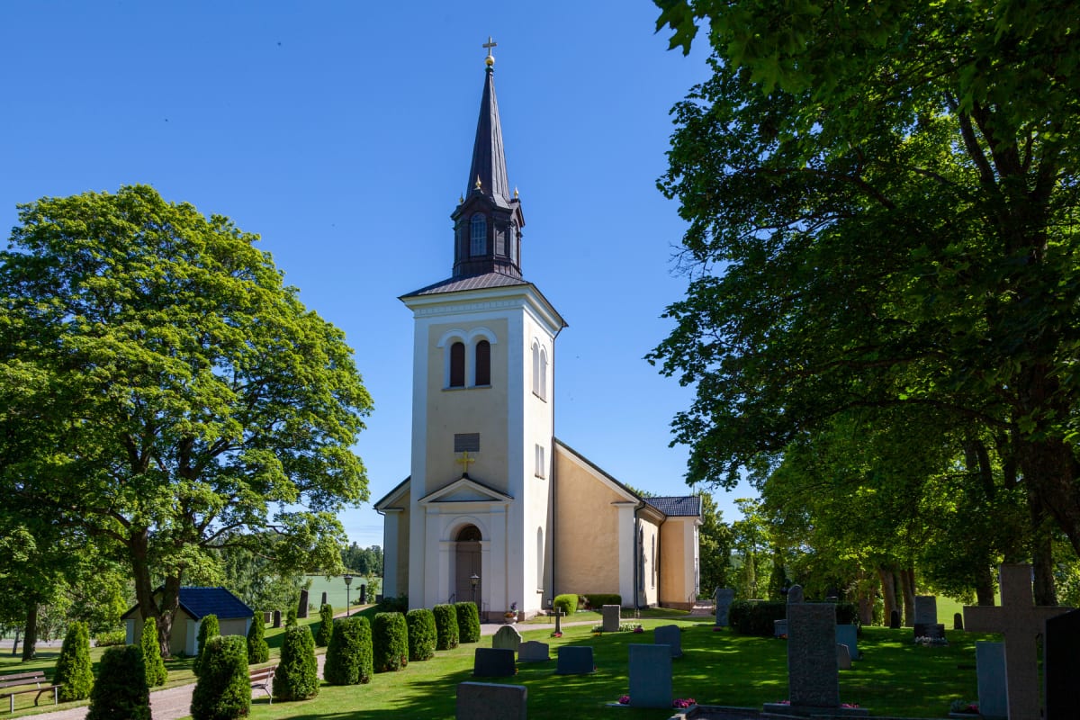 En kyrkobyggnad med ett högt torn och spetsig spira står i centrum av bilden, omgiven av en kyrkogård med gröna gräsmattor och gravstenar. Stora gröna träd ramar in kyrkan på båda sidor under en klarblå himmel.