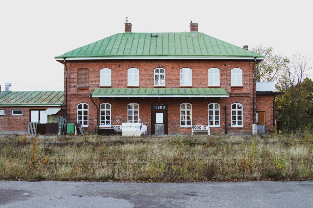 En fasadbild på en tegelbyggnad med grönt tak och vita fönster. Framför byggnaden finns ett område med torrt gräs och buskar, samt en asfalterad yta i förgrunden. Ovanför ytterdörren står "TIBRO".