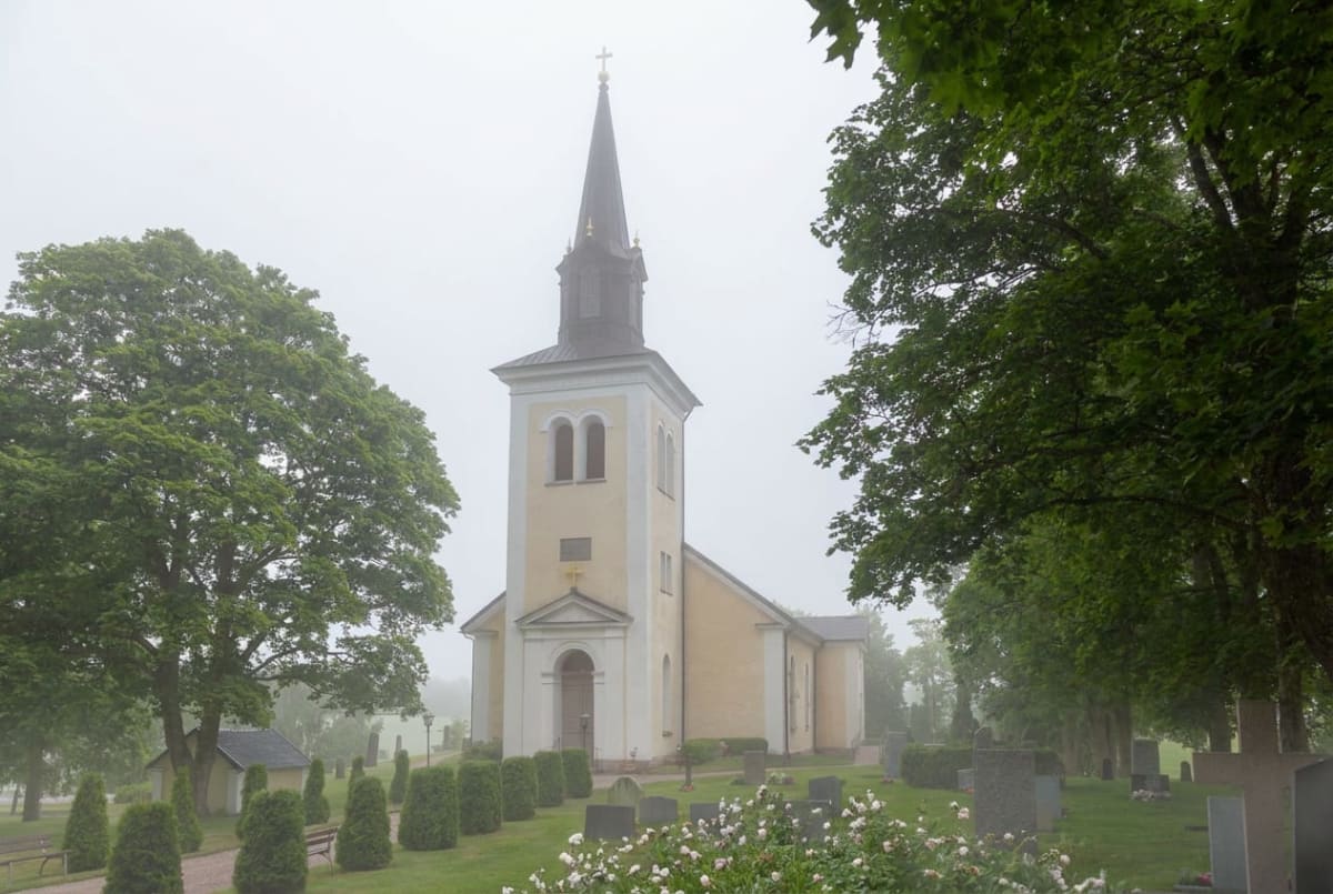 En gul kyrka med ett spetsigt koppartak och torn är centralt placerad i bilden. Runt kyrkan finns en kyrkogård med gravstenar och grönskande träd, buskar och blommor. En del av kyrkogården verkar vara omgiven av en grusgång.
