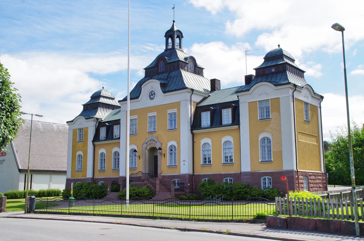 En gul byggnad med vita detaljer och ett mörkgrönt tak under en blå himmel med vita moln dominerar bilden. Framför byggnaden finns en trottoar, en gata och en svart metallstaket som ramar in en gräsmatta med buskar. Ett antal fönster med vita karmar