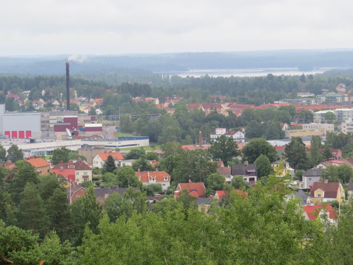 En översiktsbild av ett samhälle med bostadshus, industribyggnader, träd och i bakgrunden en sjö eller större vattensamling under en gråmulen himmel. I centrum av bilden syns en skorsten med rök som stiger uppåt och flera byggnader med röda tak.