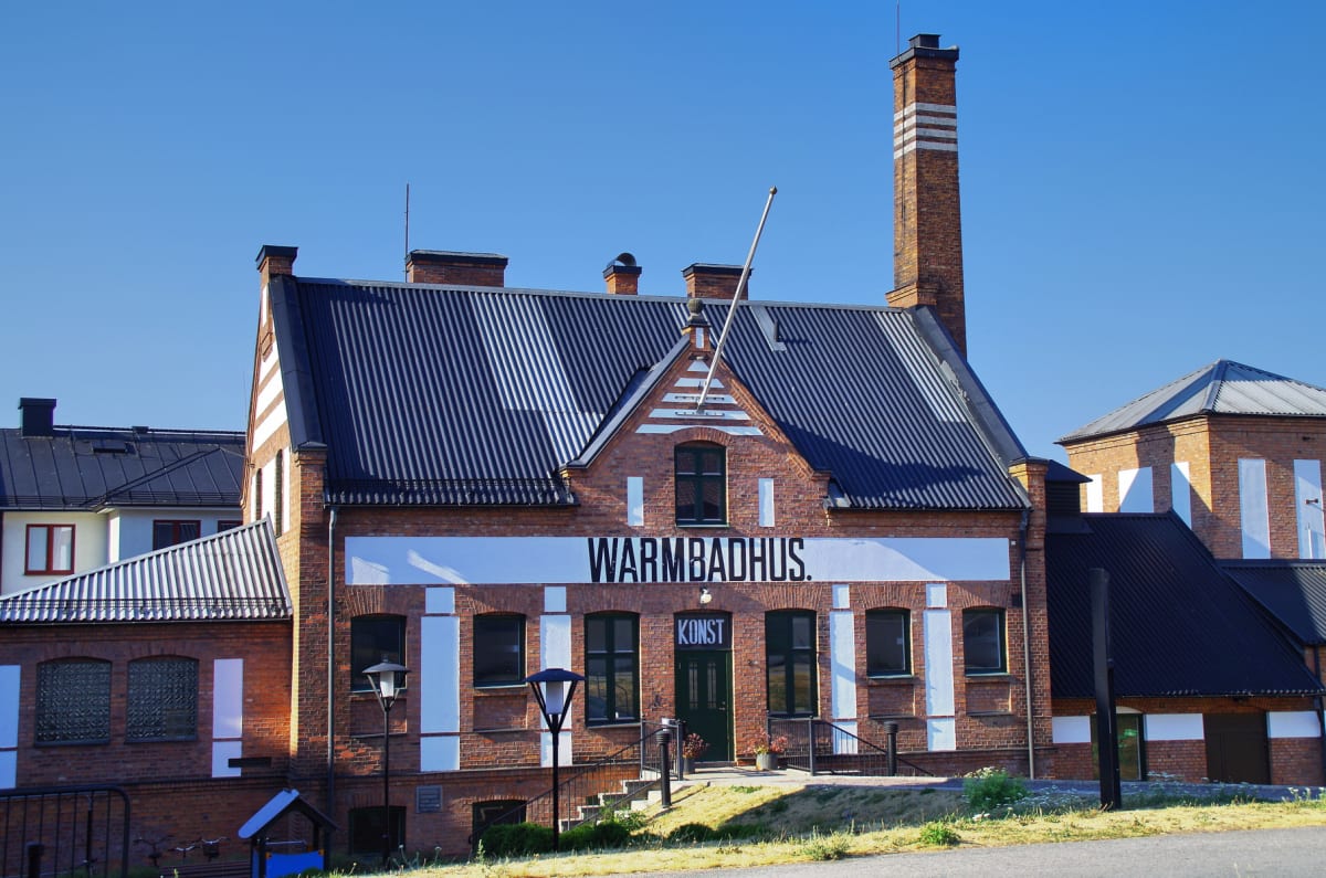 En röd tegelbyggnad vid namn "Warmbadhus" med vita detaljer och en svart plåtfasad på taket. Byggnaden har flera fönster och en grön dörr, framför vilken en trappa leder upp. På en horisontell vit banderoll står texten "WARMBADHUS." och under denna s