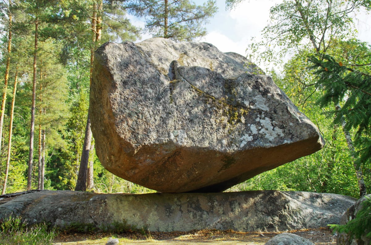 En stor, grå stenblock vilar i obalans på ett annat, plattare stenblock i en skogsmiljö. Stenarna är omgivna av gröna träd och vegetation under en klarblå himmel. Ljuset kommer från höger, skuggor är synliga under det stora stenblocket.