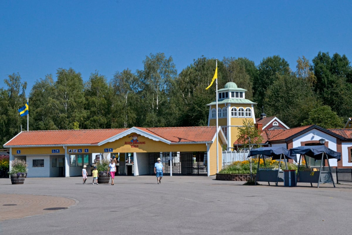 En entrébyggnad i gult med orange tak framför en trädbevuxen bakgrund under en klarblå himmel. Flera personer, inklusive barn, går mot eller bort från entrén. En svensk flagga syns till vänster och en gul vimpel på en tornbyggnad till höger. Skyltar