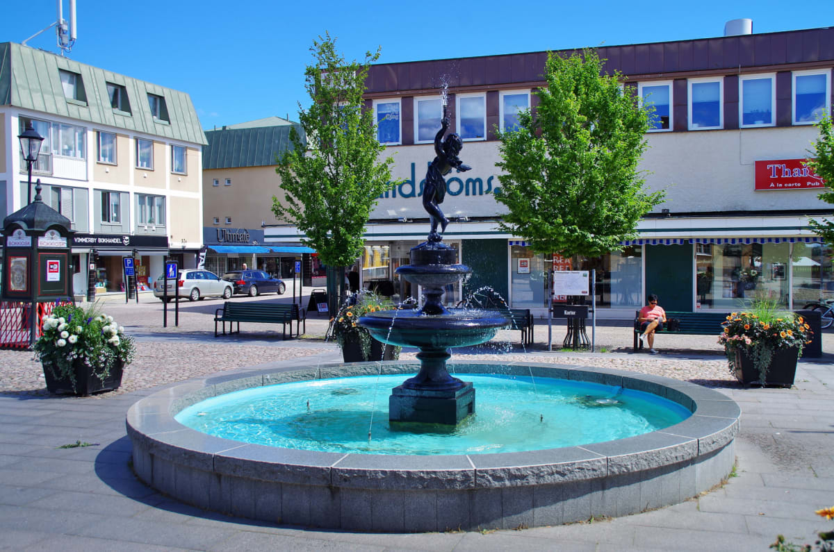 En översiktsbild av en stadsmiljö med en fontän i förgrunden, omgiven av butiker och träd. En person sitter på en bänk nära fontänen. Skyltar för "VIMMERBY BOKHANDEL" och "Thai À la carte Pub" syns tydligt.