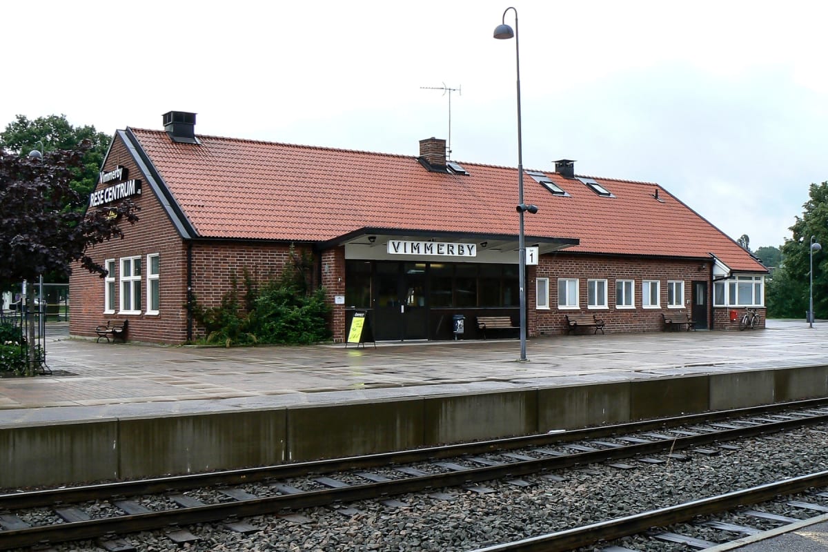 En bild som visar en tegelbyggnad med ett tegeltak och skyltar där det står "VIMMERBY RESECENTRUM". Framför byggnaden finns en perrong och tågräls. Bänkar, en lyktstolpe, cyklar och gröna buskar syns också i bilden.