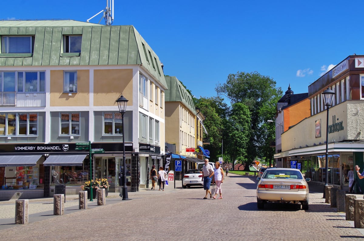 En gata i en stadsmiljö med byggnader på båda sidor, kullersten på marken, och flera personer synliga. Två bilar är parkerade eller kör på gatan. Himlen är klarblå och gröna träd syns i bakgrunden.