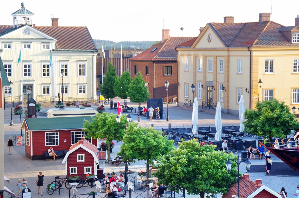 En översiktsbild av ett torg i en stad med byggnader i bakgrunden. Torget har flera träd, människor som sitter och går, kafébord med parasoller, samt två röda stugliknande byggnader i förgrunden.
