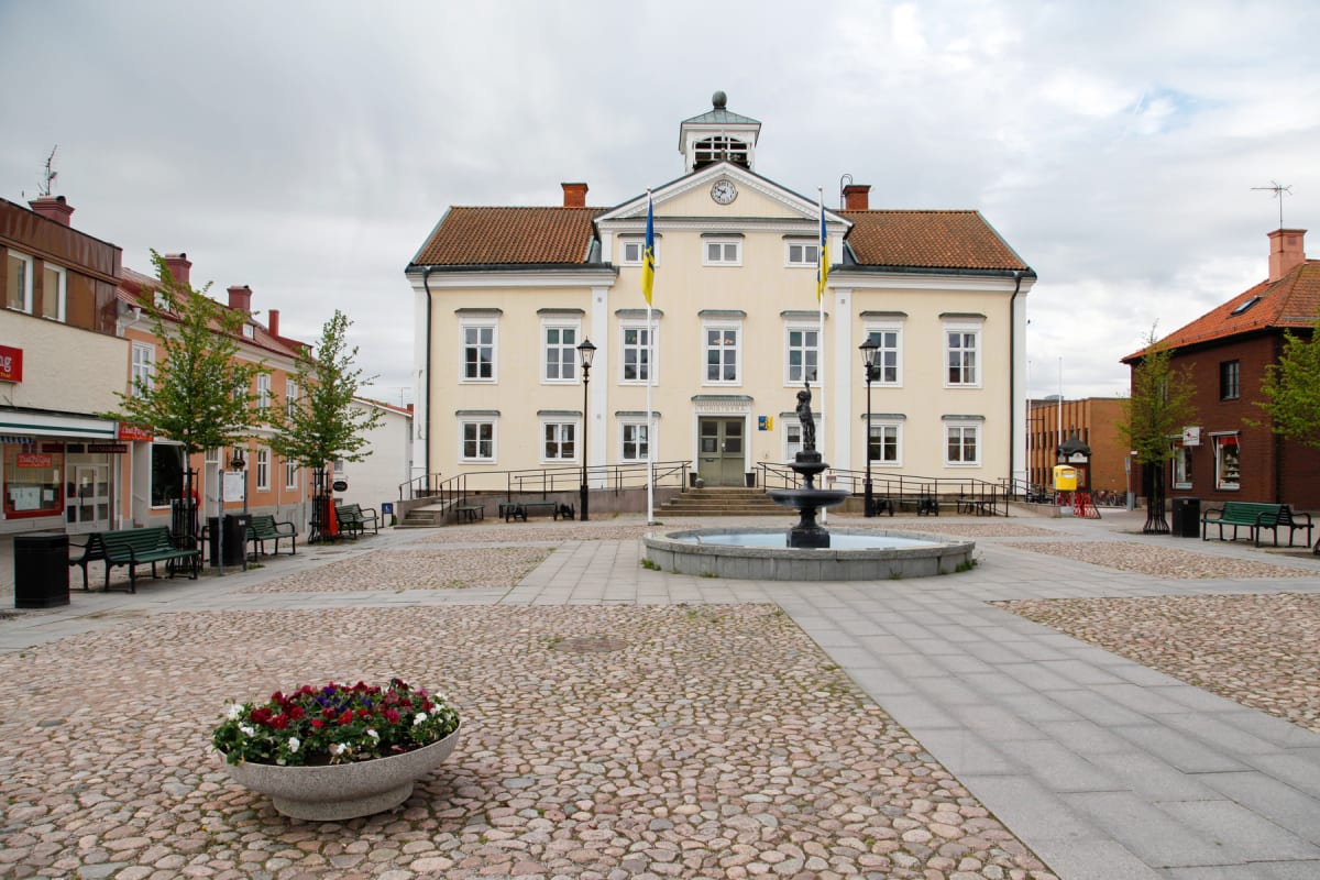 En bild som visar ett torg med en stor ljusgul byggnad i mitten. Byggnaden har ett torn med en klocka högst upp och en stor trappa leder upp till huvudentrén. Framför byggnaden finns en fontän med en staty i mitten. Torget är belagt med gatsten och h