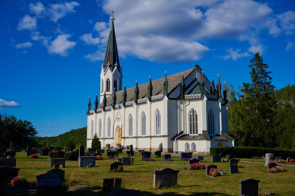 En stor vit kyrkobyggnad med spetsigt torn och många höga, smala fönster under en klarblå himmel med vita moln. Framför kyrkan syns en kyrkogård med gravstenar och blommor, omgiven av grönska och träd. Fjärran synes en grönklädd skog. Kyrkans tak har