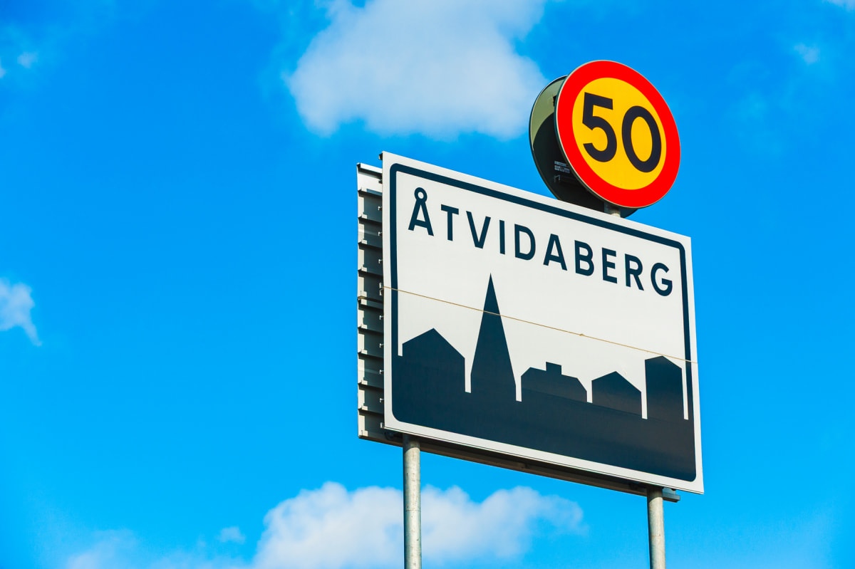En vägskylt med texten ”ÅTVIDABERG” och en grafisk silhuett av byggnader, samt en hastighetsbegränsningsskylt på 50 km/h, mot en klarblå himmel med några vita moln i bakgrunden.