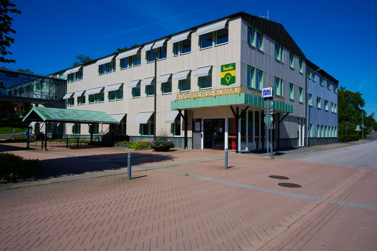 En stor, trevåningsbyggnad med ljusa fasader och många fönster med ljusgröna karmar och grå markiser. På fasaden finns en stor skylt med texten "Åtvidabergs kommun" och en kommunlogotyp. Framför byggnaden finns en markyta belagd med rödaktiga stenpla