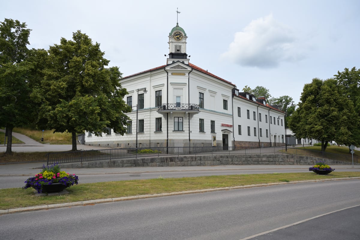En vit byggnad med ett klocktorn syns i mitten av bilden, omgiven av gröna träd och en väg. På byggnaden står det "KULTURHUSET" i röd text ovanför en dörr och "ADELSKÖLD" under klocktornet. Två stora blomkrukor med färgglada blommor står vid vägkante