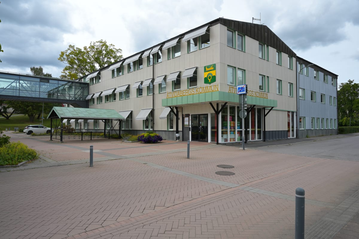 En stor byggnad med beige och ljusblå fasad. En grön entréskylt med texten "Åtvidabergs Kommun" är placerad ovanför huvudentrén. Flera fönster med vita solskydd syns längs fasaden. En inglasad gångbro förbinder byggnaden med en annan del strukturen.