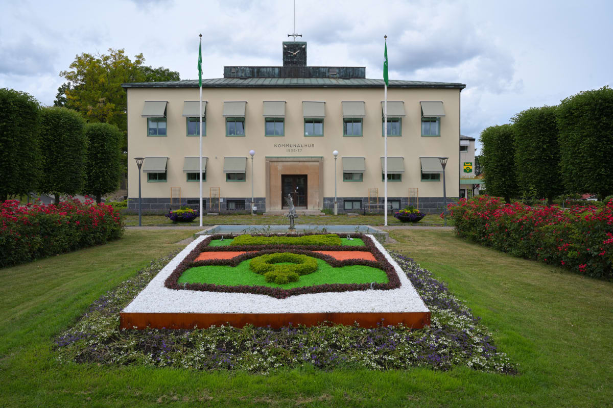 En beige byggnad med texten "KOMMUNALHUS 1936-37" ovanför entrén, flankerad av två flaggstänger med gröna flaggor. Framför byggnaden finns en gräsmatta med en mönstrad plantering i vitt, grönt och orange, samt röda buskar längs sidorna. En staty och