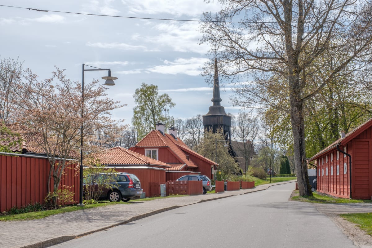 En väg kantad av röda trähus med tegeltak. Till vänster syns en parkerad personbil och en blommande buske. I bakgrunden reser sig en kyrkas spira. Träd, några med synliga knoppar och blad, ramar in bilden under en ljusblå, molnig himmel.