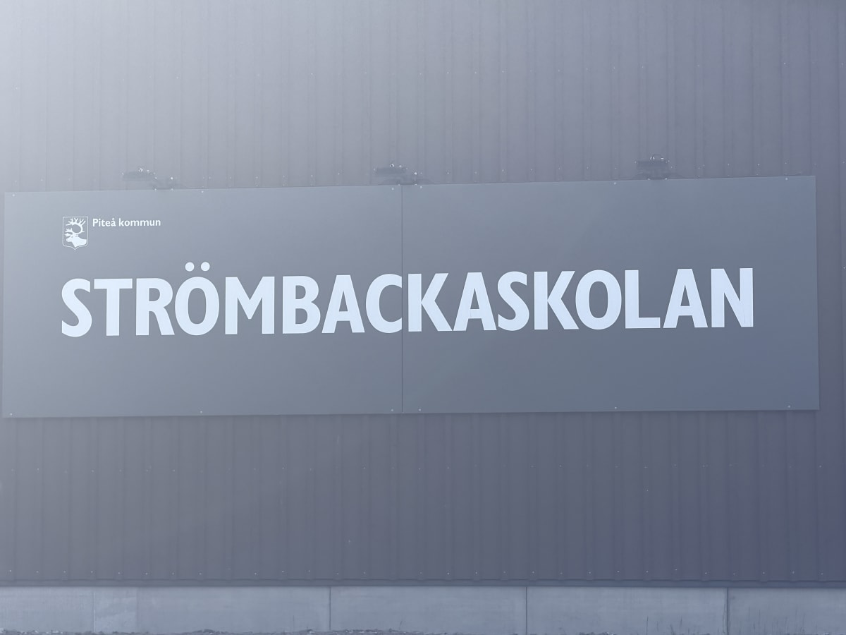 En stor, grå skylt med vit text som läser "STRÖMBACKASKOLAN" monterad på en byggnadsfasad. Ovanför texten syns Piteå kommuns logotyp och namn. Fasaden är täckt med vertikala metallpaneler, och under skylten syns en ljusare sockel.