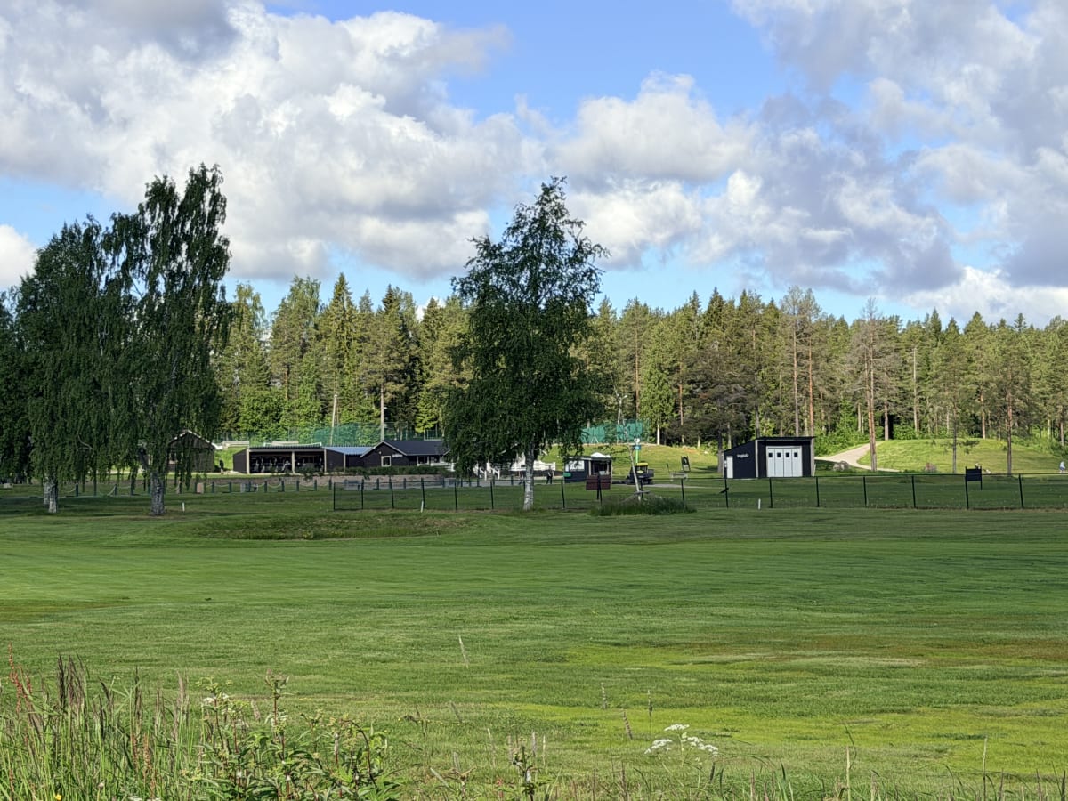 En utomhusbild som visar ett stort gräsfält med en skogskant i bakgrunden. I mitten av bilden finns några byggnader och ett staket. Två lövträd står framför skogen och himlen är blå med vita moln.