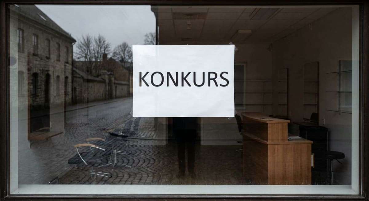 En "KONKURS"-skylt är tejpad på insidan av ett skyltfönster. Genom fönstret syns interiören med en kontorsdisk och tomma hyllor till höger, samt en kullerstensgata och stenhus i bakgrunden. Fönstret reflekterar delar av omgivningen.
