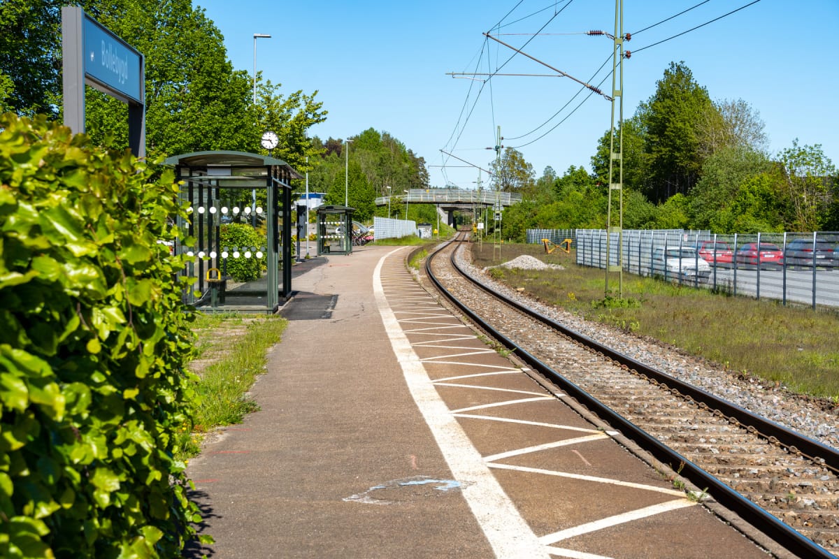 En bild som visar en tågperrong och järnvägsräls under en solig dag. Till vänster syns grönska och ett stationsskylt med namnet "Bollebygd". Perrongen har vita markeringar och i bakgrunden skymtar en bro över spåren samt parkerade bilar bakom ett sta