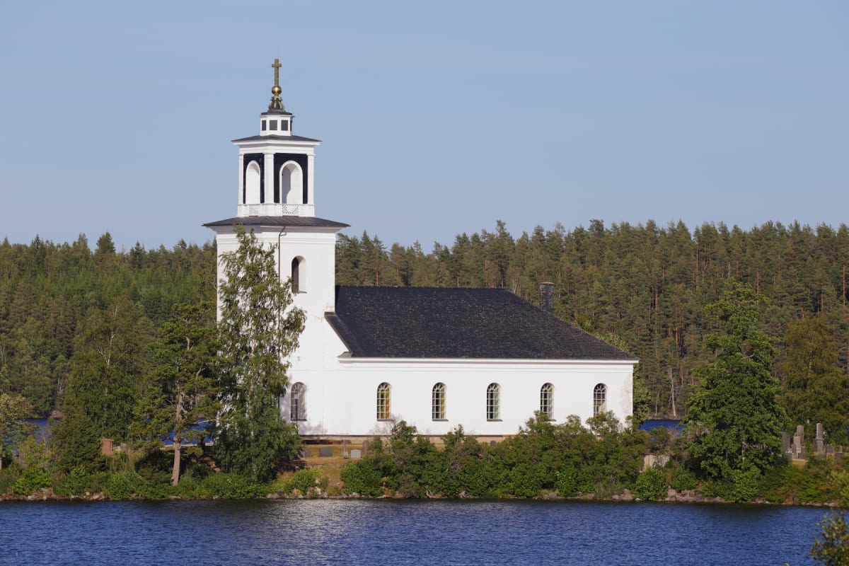 En vit kyrka med svart tak och ett torn med en gyllene kors på står vid en insjö. Bakom kyrkan syns en tät skog. Kyrkan har stora, välvda fönster längs långsidan.