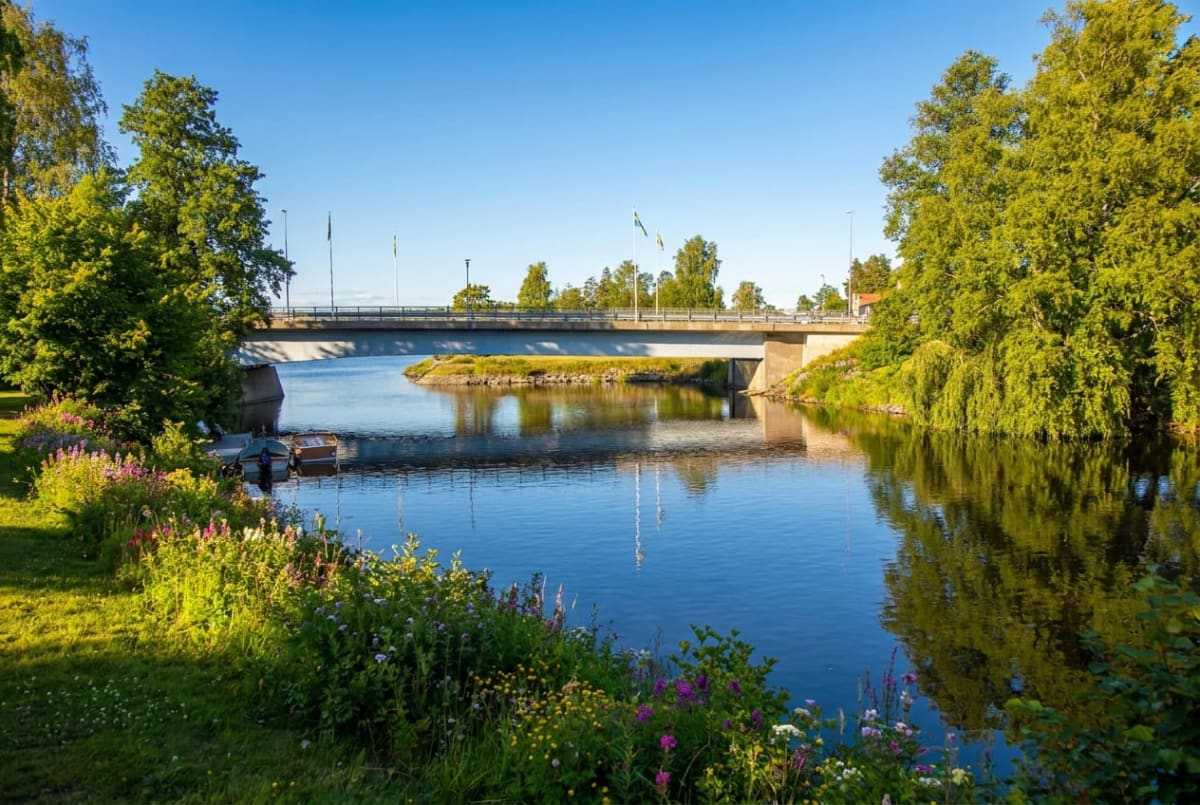 En bro sträcker sig över ett vattendrag med grönskande buskar och träd på sidorna. Två små båtar ligger förtöjda vid strandkanten till vänster. En liten ö med gräs syns under bron i mitten av vattendraget. Blå himmel ovanför.