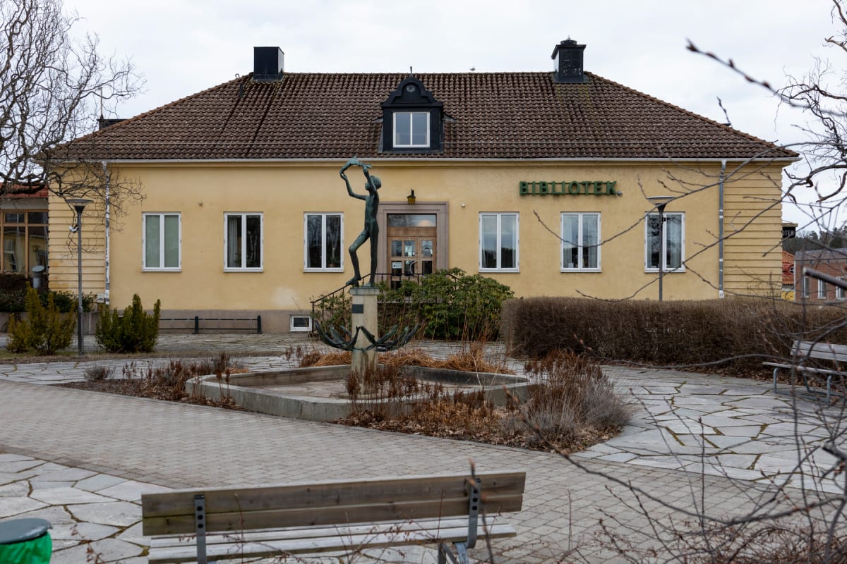 En gul byggnad med brunt tak och vita fönsterkarmar dominerar bilden. Framför byggnaden finns en bronsstaty, en fontän och en stensatt yta med sittbänkar och buskar. Ett träd utan löv syns i förgrunden till vänster.