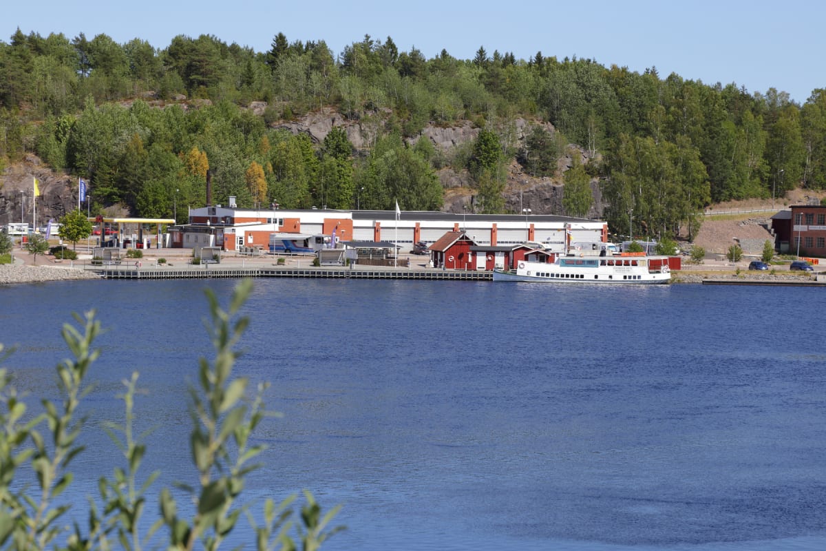 En stor byggnad med flera garageportar, en bensinstation och ett par små röda byggnader vid en brygga intill en sjö eller större vattendrag. En passagerarbåt ligger förtöjd vid bryggan. Bakom byggnaderna reser sig en trädbevuxen bergssluttning.