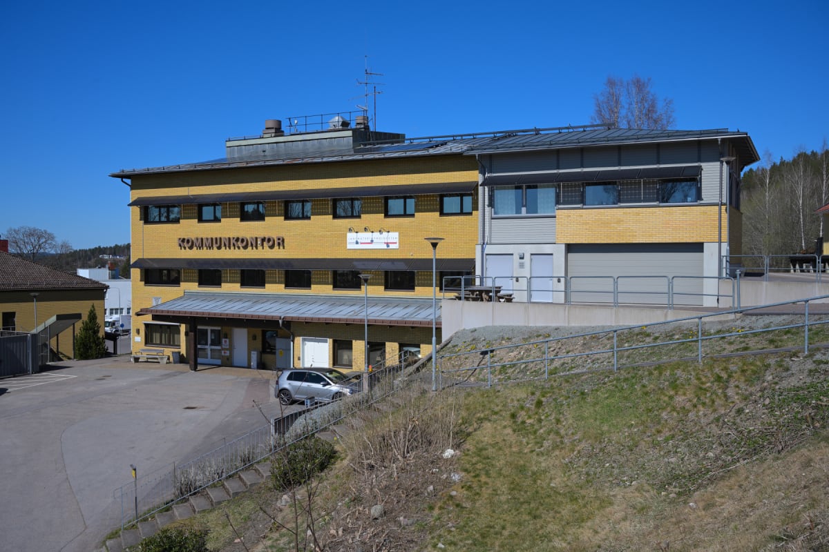 En gul tegelbyggnad med texten "KOMMUNKONTOR" upptill. En del av byggnaden har grå panel och en garageport. Framför byggnaden finns en parkeringsplats med en silverfärgad bil och en trappa med räcke som leder upp till byggnadens entré. Det är en soli