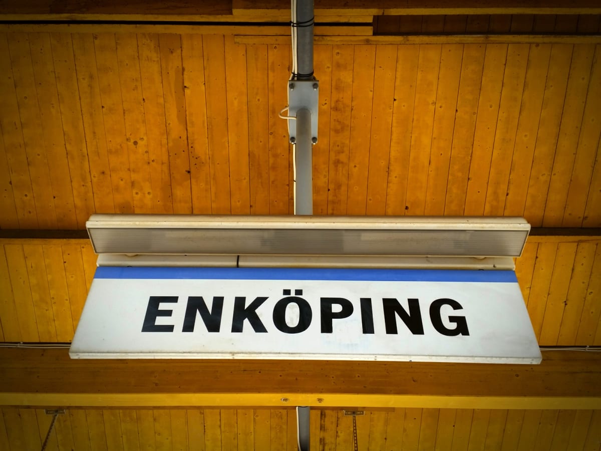 En stor, vit skylt med blå rand och texten "ENKÖPING" i svarta versaler, hänger från ett orangefärgat trätak. Ovanför skylten sitter en ljusarmatur och i taket syns en silverfärgad stolpe med kablar.