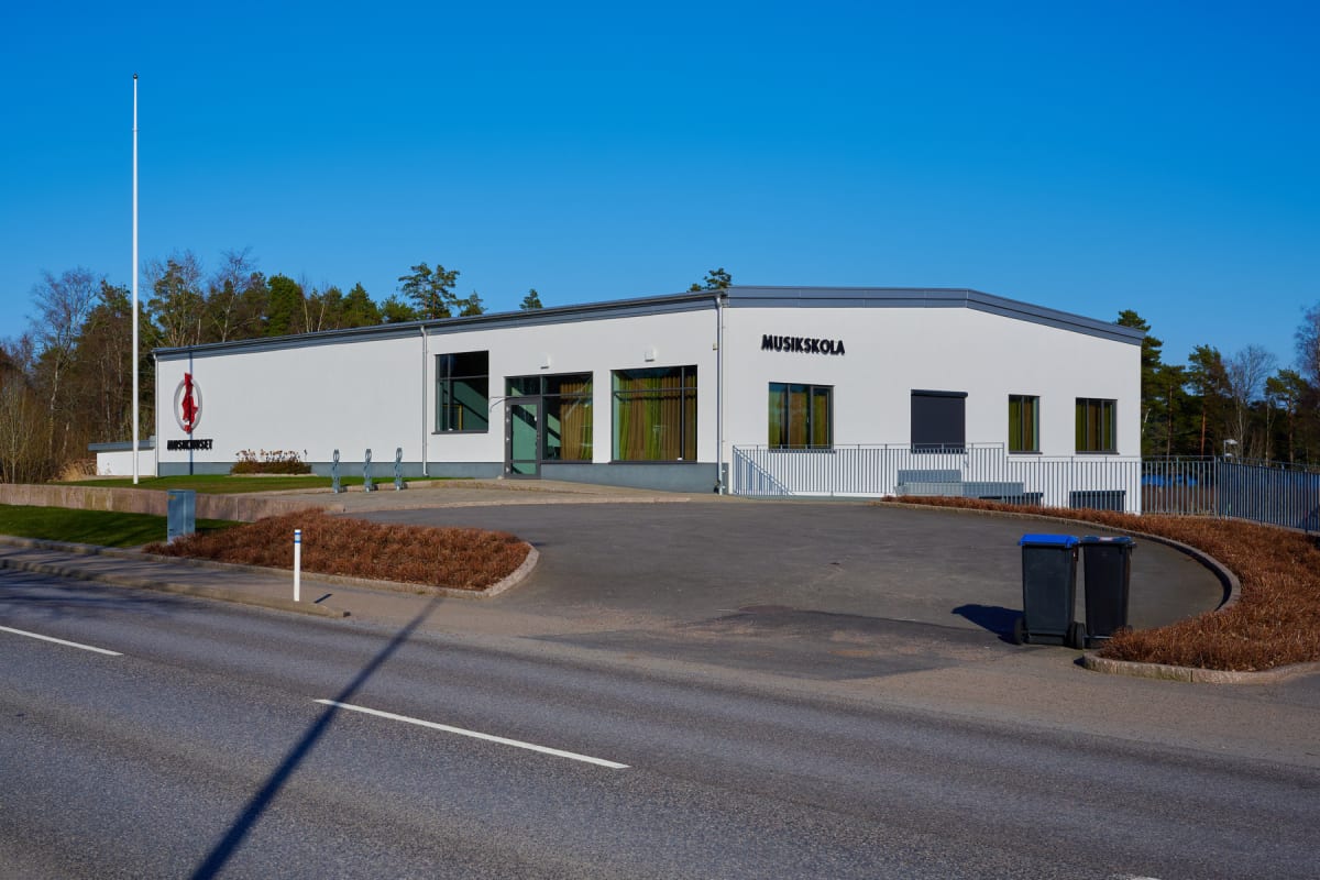 En modern vit byggnad med grå tak och fönsterramar under klarblå himmel. På byggnadens fasad står "MUSIKKOLAN" och "MUSIKHUSET" med en logotyp. Bilden visar en asfalterad yta, växtlighet, och en väg i förgrunden.