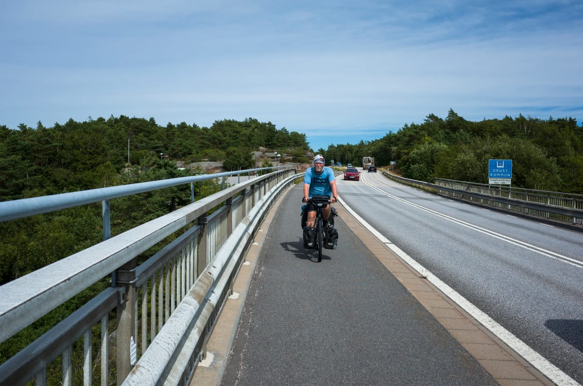 En bild som visar en person på cykel på en bro. Till vänster om cyklisten finns ett räcke och till höger en väg med bilar. Ett blått vägmärke med texten "ORUST KOMMUN" syns till höger på bilden. Omgivningen består av grönskande träd och en blå himmel