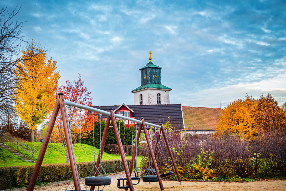 En bild visar en lekplats med gungor i förgrunden. I bakgrunden syns en kyrkobyggnad med ett grönt torn och ett tak med tegelpannor, samt hus med röda och mörkbruna tak. Lövfällande träd med gula och röda löv indikerar höst.