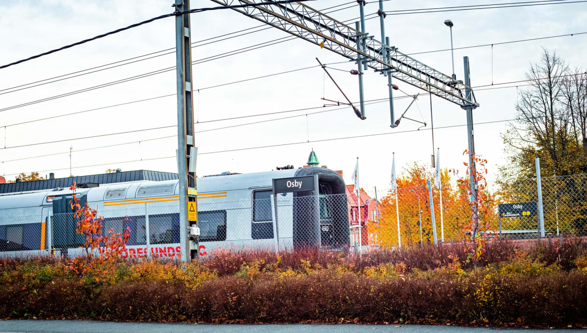 En silverfärgad tåg med gula detaljer passerar en station som heter Osby. Runt tåget finns buskar och träd med höstfärger i orange och gult. Elledningar och stolpar korsar bilden ovanför tåget.