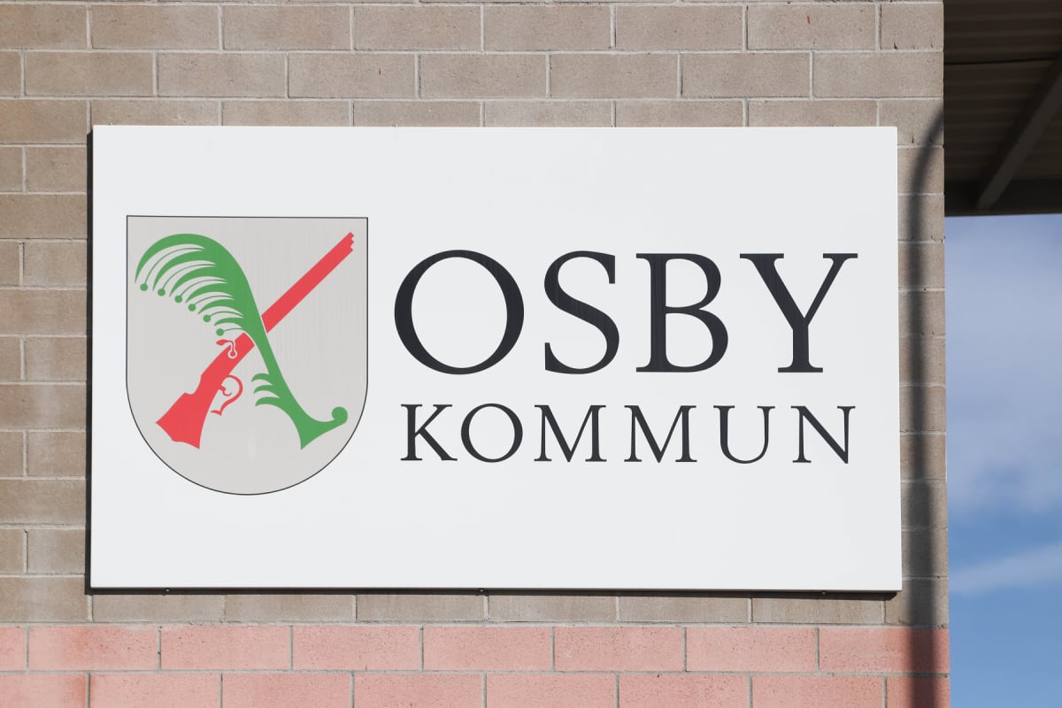 En rektangulär vit skylt med texten "OSBY KOMMUN" i svarta versaler. Till vänster på skylten finns Osby kommuns vapen, som består av två korsade gevär, ett rött och ett grönt, mot en ljusgrå bakgrund, med en grön fjäderliknande figur. Skylten är mont
