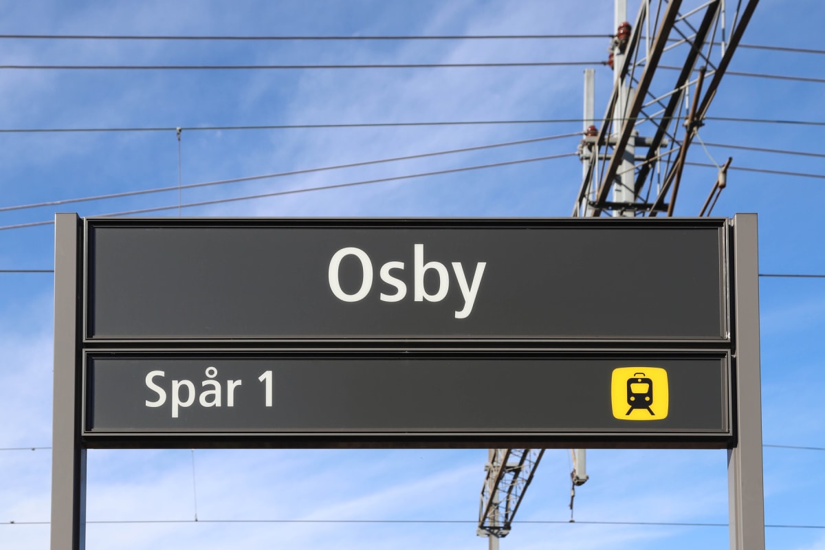 En svart stationsskylt med vit text "Osby" och "Spår 1" med en gul tågikon, under en blå himmel med tunna vita moln. I bakgrunden syns kraftledningar och metallkonstruktioner för tågtrafik.