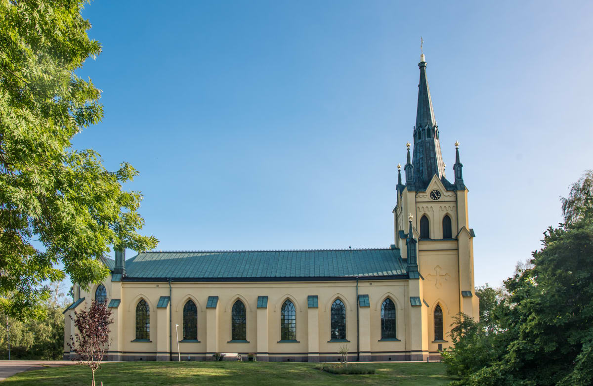 En gul kyrkobyggnad med ett högt, spetsigt torn syns under en klarblå himmel. Till vänster i bilden växer ett lummigt träd och grönskande buskar ramar in kyrkan. Markytan består av gräsmatta framför byggnaden och träd i bakgrunden.