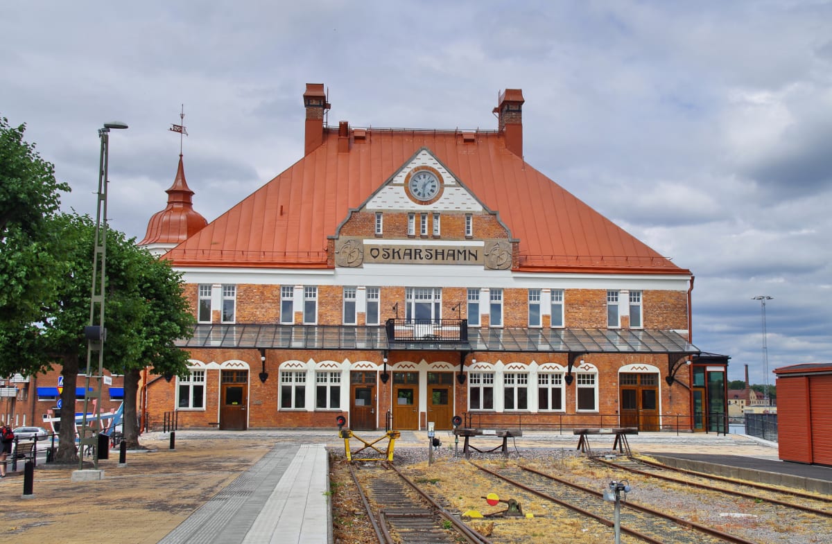 En stor, röd tegelbyggnad med ett orange tak och ett klocktorn, som ser ut att vara en gammal tågstation. Framför byggnaden finns tågspår med torrt gräs, och till vänster om spåren en asfalterad plattform. Till vänster i bilden syns en rad träd.